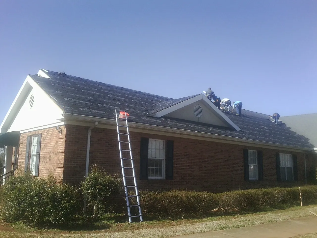Crew tearing shingles off a brick ranch roof with a ladder set — Emergency Roof Repair work by our crew in Mancelona