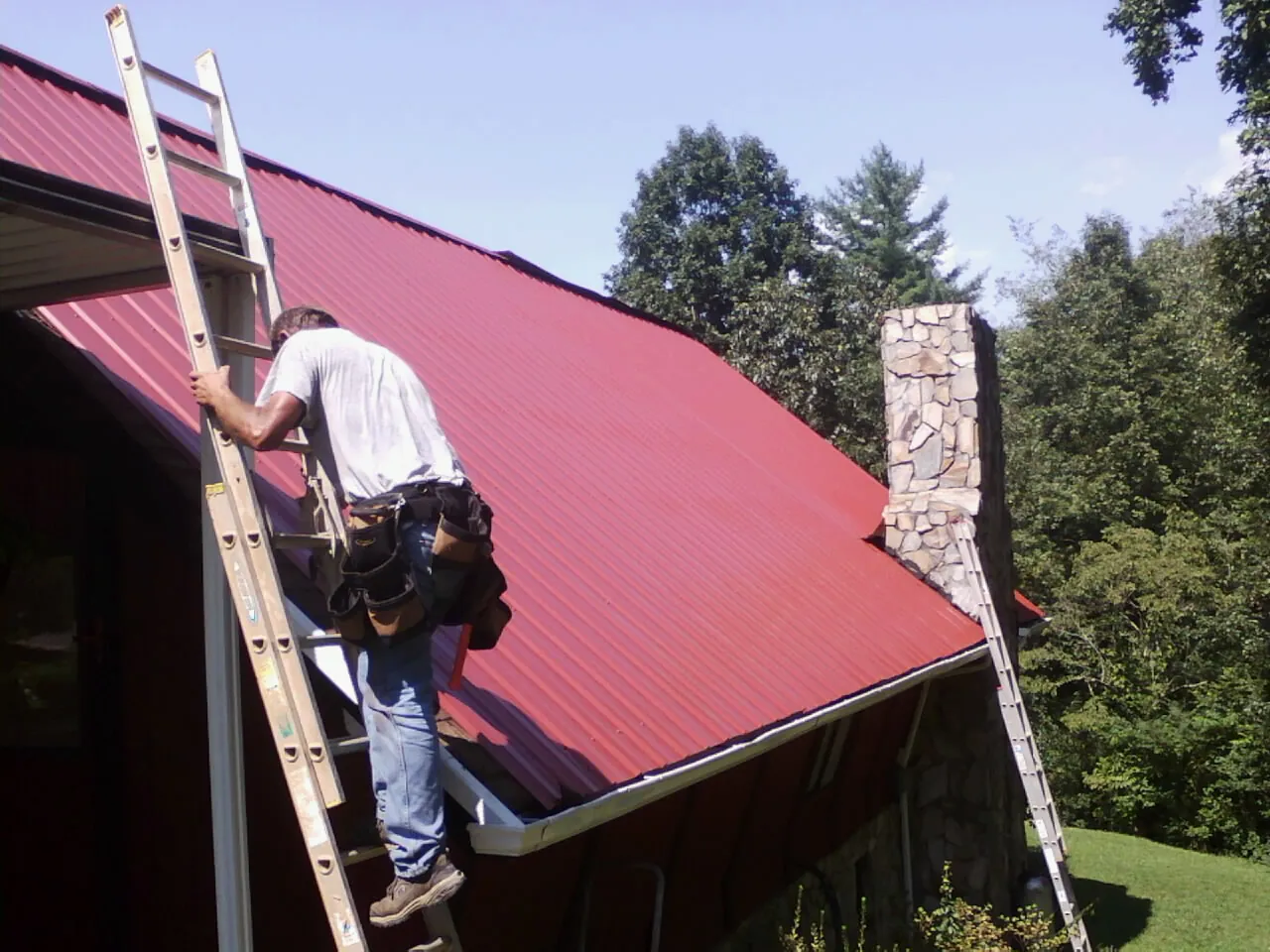 Roofer on a ladder beside a red metal roof and stone chimney — Soffit & Fascia Repair by Roofing Brothers in Mancelona
